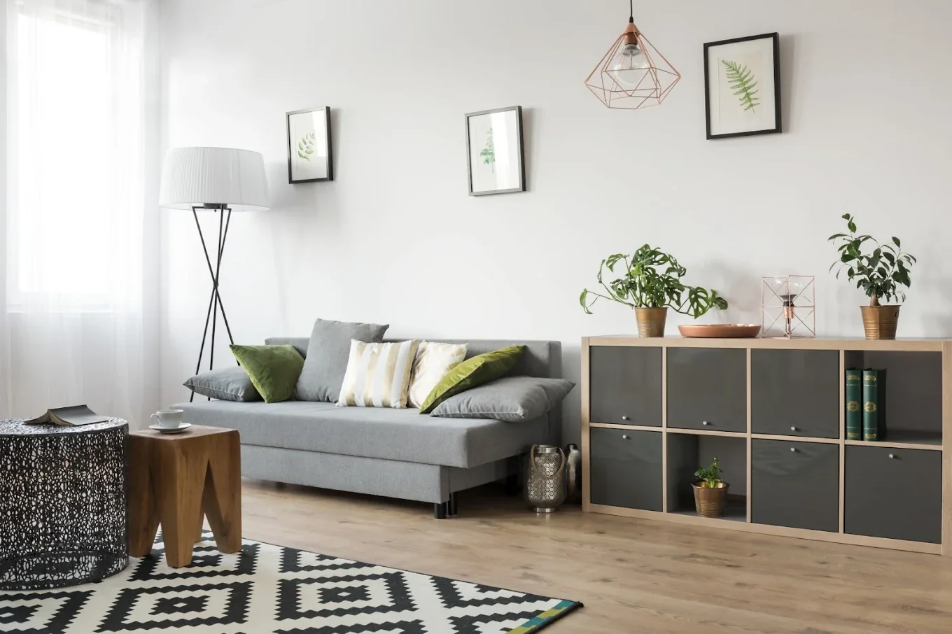 Jasny salon z szarą kanapą, zielonymi poduszkami, roślinami i drewnianymi akcentami